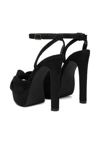 Černé platformové podpatky se semišovou texturou, s uzlovým designem vpředu a štíhlým páskem kolem kotníku se sponou. Vysoký jehlový podpatek.