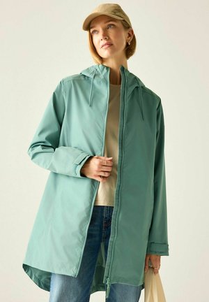 Frau mit beiger Kappe, türkisfarbener Kapuzen-Regenjacke über beigem Hemd, blauen Jeans, hält eine beige Tragetasche, blickt leicht nach oben.