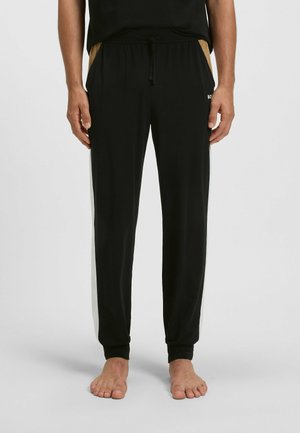 Homme portant un pantalon de jogging noir avec des rayures blanches sur les côtés et des accents de poche beige, debout pieds nus contre un fond uni.