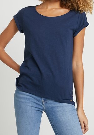 Frau trägt ein marineblaues T-Shirt mit kurzen Ärmeln und hellblaue Jeans, steht mit einer Hand in der Hüfte vor einem einfarbigen Hintergrund.