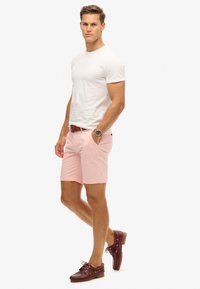 Camiseta blanca de cuello redondo, pantalones cortos rosa claro, cinturón marrón, zapatos barco marrones. El modelo posa con las manos en los pockets, mostrando un atuendo casual de verano.