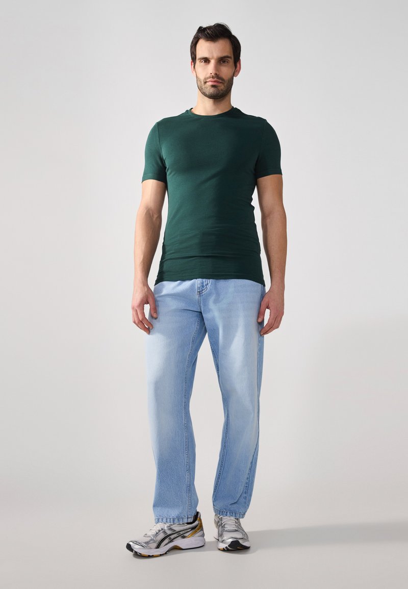 Grønn kortermet skjorte kombinert med lyseblå denimjeans og sølvfargede joggesko. Enkelt design, tettsittende skjorte og avslappet passform på jeansene.