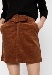 Bruine corduroy rok met een gestructureerd oppervlak, uitgerust met een bijpassende riem en voorzakken, afgeknipt boven de knie. Inclusief riemlussen.