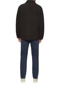 Schwarze Hemdjacke aus einer Wollmischung mit Kragen, langen Ärmeln und Knopfdetails, kombiniert mit dunkeltblauen Jeans und hellen Schuhen.