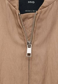 Bež zip-up majica s visokim ovratnikom, vertikalnom rebrastom teksturom i srebrnim metalnim patentnim zatvaračem sprijeda.