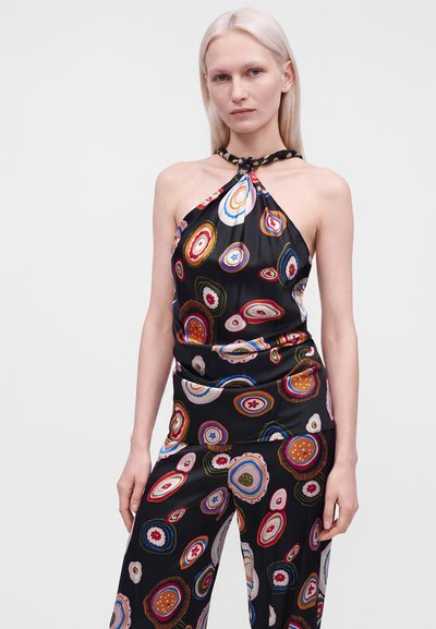 Femme aux cheveux blonds clairs et raides portant un haut à bretelles noir et un pantalon avec un motif circulaire coloré sur un fond uni.