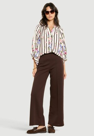 Femme portant une blouse fleurie rayée, un pantalon large foncé, des lunettes de soleil et des mocassins marron, debout devant un fond clair uni.