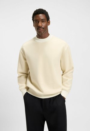 Krämfärgad sweatshirt med hög hals och ribbade ärmslut, tillverkad av mjukt tyg. Bärs med svarta, avslappnade byxor.