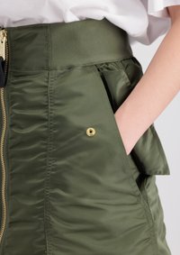 Gonna puffer verde oliva con una ampia fascia elastica in vita, chiusura frontale con zip e tasca laterale con dettaglio di bottone a pressione dorato.