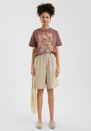 Braunes Grafik-T-Shirt mit floralem Design, beige Shorts, cremefarbene Espadrilles und eine Mesh-Umhängetasche. Das Modell steht aufrecht vor einem hellen Hintergrund.