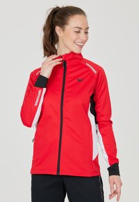 Giacca sportiva rossa con colletto alto, dettagli in bianco e nero, tessuto testurizzato e particolari riflettenti. Chiusura con zip frontale e tasche laterali con zip.