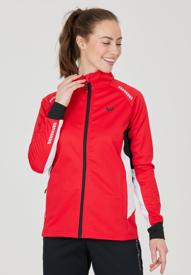 Giacca sportiva rossa con colletto alto, dettagli in bianco e nero, tessuto testurizzato e particolari riflettenti. Chiusura con zip frontale e tasche laterali con zip.