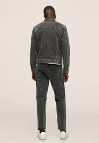 Uomo che indossa una giacca di jeans grigio scuro e jeans, in piedi con le spalle alla telecamera su uno sfondo chiaro e uniforme.