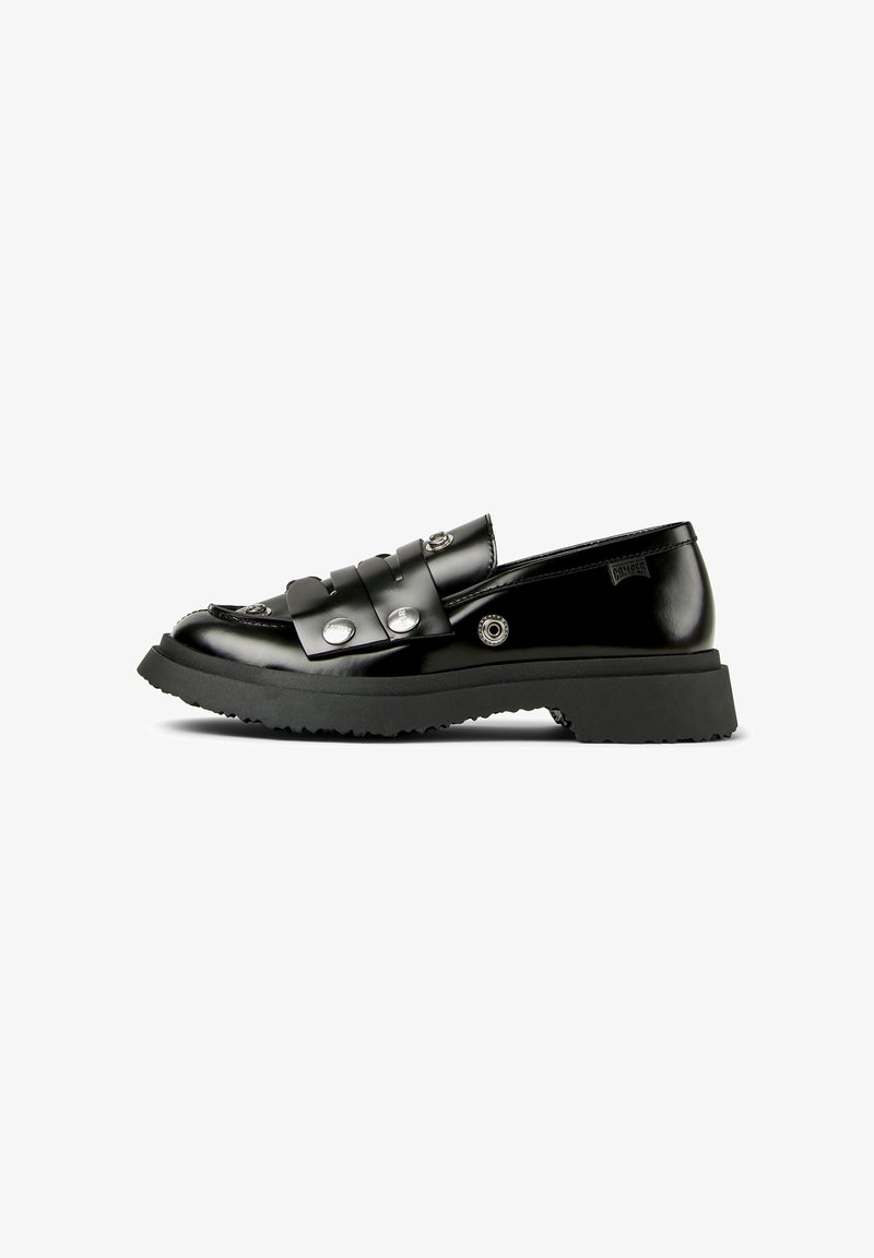 Mocassin en cuir noir brillant avec semelle en caoutchouc épaisse, détails à clous argentés, et une bride sur le dessus, présenté sur un fond blanc.