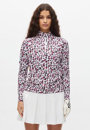 Frau trägt ein langärmliges, pink-schwarz-weißes, leopardgemustertes Jackett mit Reißverschluss, weißen plissierten Rock und einen weißen Golfhandschuh an der rechten Hand.