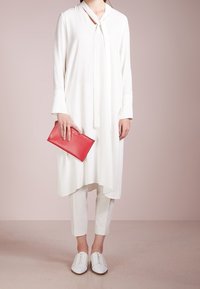 Túnica blanca de manga larga con cuello atado, acompañada de pantalones blancos ajustados y zapatos blancos sin cordones. Sosteniendo un bolso de mano rojo.