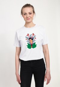 Bílý bavlněný tričko s barevným květinovým a portrétním motivem, se zelenými listy a červeným srdcem. Model nosí černé kalhoty.
