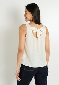 Haut sans manches beige clair avec une finition texturée, présentant un dos en trou de serrure et un accent de nœud central. Porté avec un jean foncé.