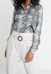 Camicia a maniche lunghe in stampa serpente bianco e nero, con chiusura a bottoni davanti, abbinata a pantaloni beige chiaro a vita alta con un cinturino circolare.