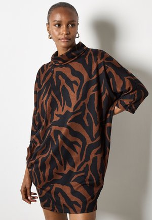 Donna che indossa un vestito oversize a strisce tigre marroni e nere con un collo alto e maniche a tre quarti, in piedi contro un muro bianco.