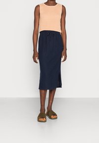 Top sans manches beige à côtes, jupe midi marine avec taille élastique et fente sur le côté, associée à des sandales croisées vert olive avec semelles beiges.