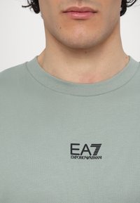 T-shirt verde chiaro a maniche corte realizzato in morbido cotone con colletto a girocollo a coste. Presenta un logo EA7 Emporio Armani nero sul petto.