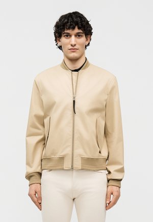 Νεαρός άνδρας με κατσαρά μαλλιά φοράει μια μπεζ μπουφάν τύπου bomber με φερμουάρ, στέκεται μπροστά σε ένα απλό φωτεινό φόντο, κοιτάζοντας προς τα εμπρός.