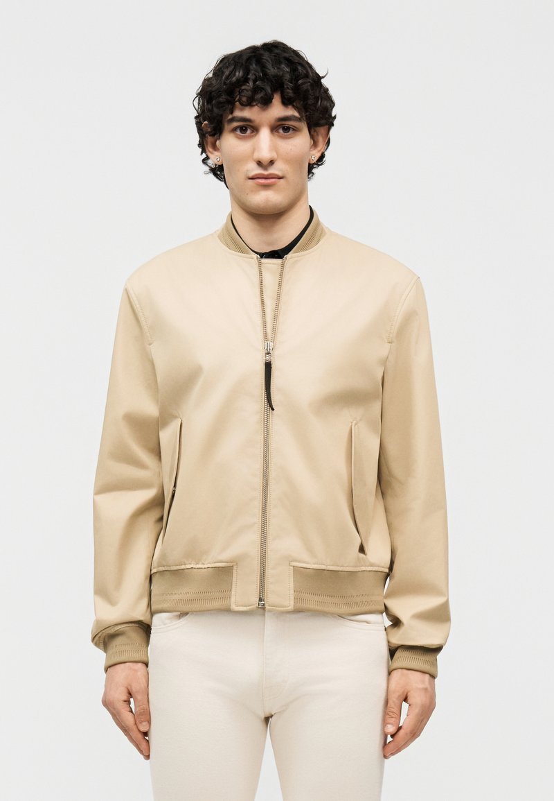 Göndör hajú fiatal férfi bezipzározott bézs bomber dzsekit visel, sima világos háttér előtt áll, előre nézve.