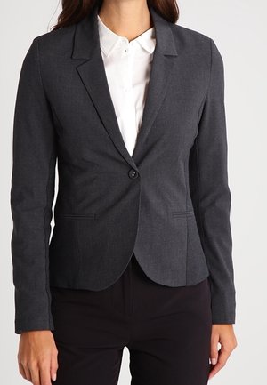 Vrouw draagt een aansluitende donkergrijze blazer over een wit overhemd en zwarte broek, zichtbaar van nek tot heupen.