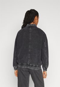 Black denim jacket with a relaxed fit, featuring a collar, elastic cuffs, and a hem. The fabric has a faded texture and visible stitching.