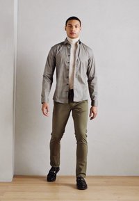 Camisa de botones en un patrón de cuadros gris y blanco, combinada con pantalones verdes oliva y zapatos negros. Tejido suave, corte relajado, estilo casual.
