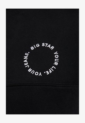 Valge tikandiga tekst "BIG STAR YOUR LIFE, YOUR JEANS" mustal kangal ringis, horisontaalse õmbluse kohal.