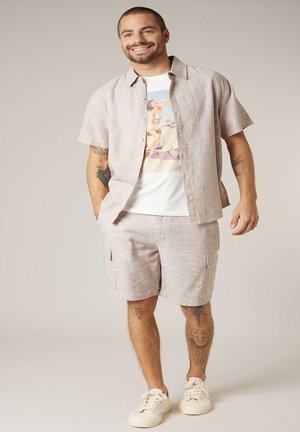 Homme souriant portant une chemise à manches courtes beige boutonnée et un short assorti, t-shirt graphique, baskets blanches, debout devant un fond neutre.