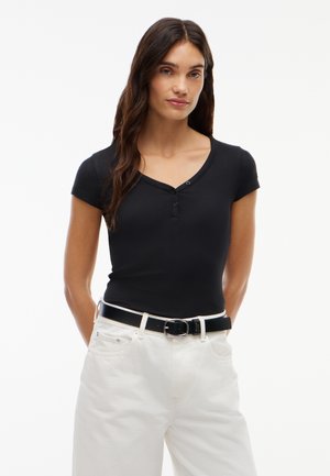 Femme aux longs cheveux bruns portant un haut noir boutonné à manches courtes, un pantalon blanc et une ceinture noire sur fond blanc.