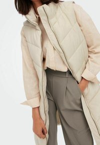 Kobieta w beżowej, pikowanej kamizelce bez rękawów, pod którą ma jasnobeżową koszulę na guziki i szare spodnie z wysokim stanem, z ręką w kieszeni.