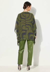 Gennemsigtig, mønstret olivengrøn top med mørke geometriske designs, parret med grønne, glansfulde, tapered bukser og metalliske sølvhæle.