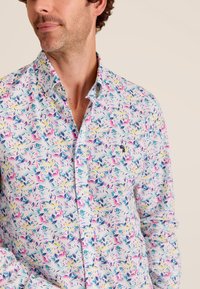 Bloemige button-up shirt met een lichte stof, een witte basis en levendige patronen van roze, geel en blauwe bladeren, en een klein logo op de borst.