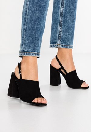 Fødder iført sorte ruskind åbne højhælede sandaler med ankelremme, kombineret med blå denimjeans med opsmøgede ben over anklerne.