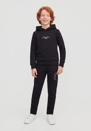 Enfant roux portant un sweat à capuche noir et un pantalon de jogging assorti avec des logos blancs en écriture, debout et souriant sur un fond gris uni.