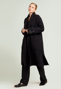 Lange zwarte wollen jas met grote zakken en een sjaalkraag. De manchetten zijn versierd met ingewikkelde kralenpatronen. Draagt deze over zwarte broek en loafers.