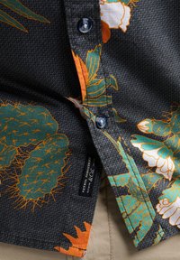 Chemise à manches courtes en tissu texturé foncé avec un motif de cactus et de fleurs en vert, orange et blanc, boutons contrastants et étiquette.