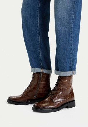 Dunkelbraune Stiefeletten aus Leder mit Krokodilprägung, Seitenreißverschluss und Schnürsenkeln, getragen zu hochgekrempelten blauen Jeans, vor hellem Hintergrund.