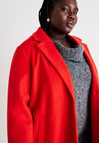 Rød ullfrakk med en strukturert krage, som har to sidelommer og en myk, grå strikket genser under med cowl neck-design.