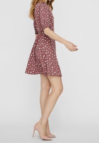 Frau in einem mauvefarbenen Kurzarmkleid mit weißen Punkten, die seitlich vor einem schlichten Hintergrund steht, dazu nude farbige High Heels.