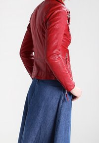Red leather jacket with a fitted design, asymmetrical zipper, and textured finish. Worn with a blue denim skirt featuring a side zipper.