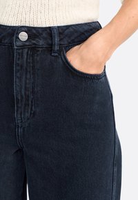 Jean en denim bleu foncé avec une taille haute, coupe droite, poche latérale visible et détails en bouton argenté. Le tissu semble texturé et durable.