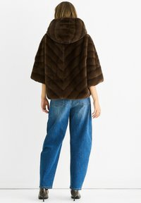 Woman wearing a brown fur hooded jacket with 3/4 sleeves, blue loose jeans, and pointed high-heeled boots standing against a white background.