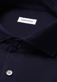 Colletto della camicia blu navy con un'etichetta in tessuto bianco con la scritta "seidensticker" e un bottone nero con quattro fori sottostante. Texture liscia.