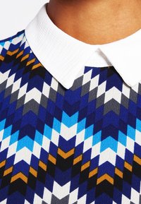 Suéter de punto con un patrón en chevron azul, blanco y negro, con un cuello blanco texturizado y fuertes contrastes de color geométricos.