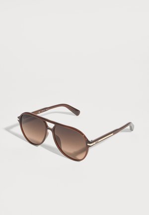 Gafas de sol aviador marrones con lentes marrones degradadas y finos detalles dorados en las patillas, colocadas sobre una superficie blanca lisa.